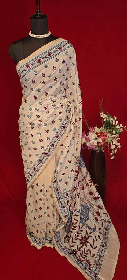 Handcrafted Kantha stitched silk blend saree in cream with multicolor floral and bird embroidery in blue tones on a mannequin with flower vase against a red background.