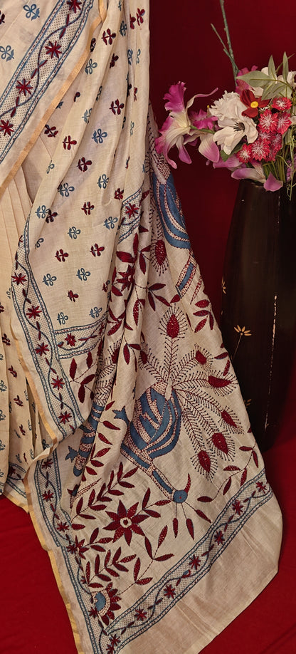 Handcrafted Kantha stitched silk blend saree in cream with multicolor floral and bird embroidery in blue tones on a mannequin showcasing saree pallu with flower vase against a red background.