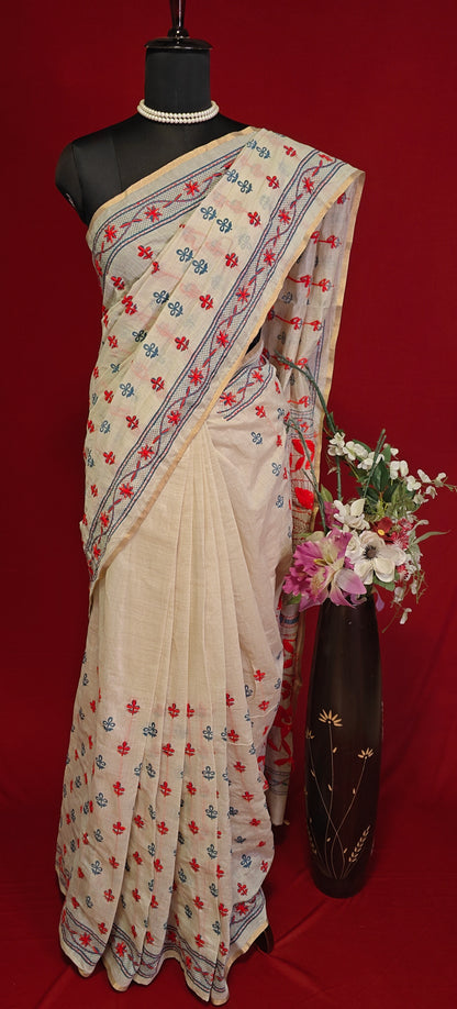 Handcrafted Kantha stitched silk blend saree in cream with multicolor floral and bird embroidery on a mannequin along with a flower vase against a red background.