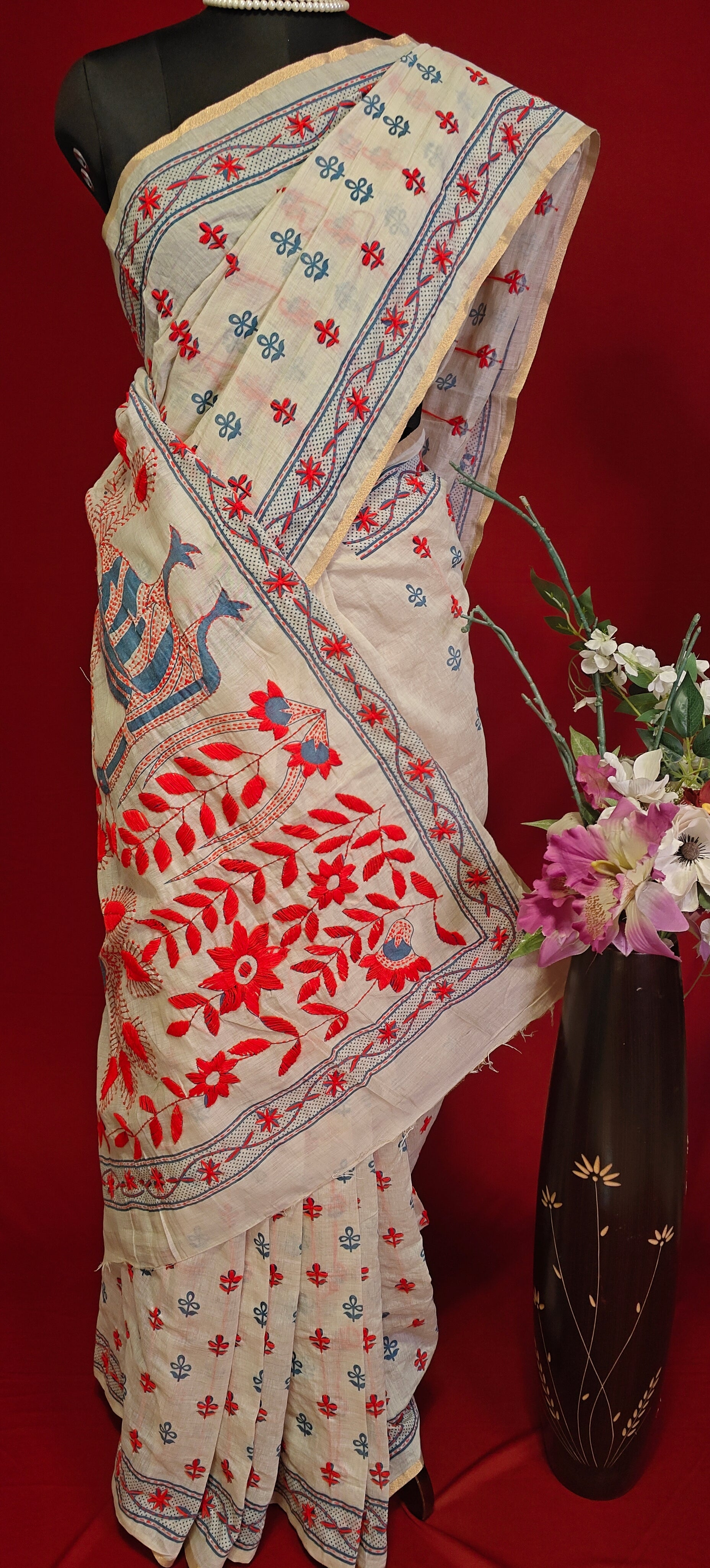 Handcrafted Kantha stitched silk blend saree in cream with multicolor floral and bird embroidery on a mannequin showcasing the saree pallu along with a flower vase against a red background.