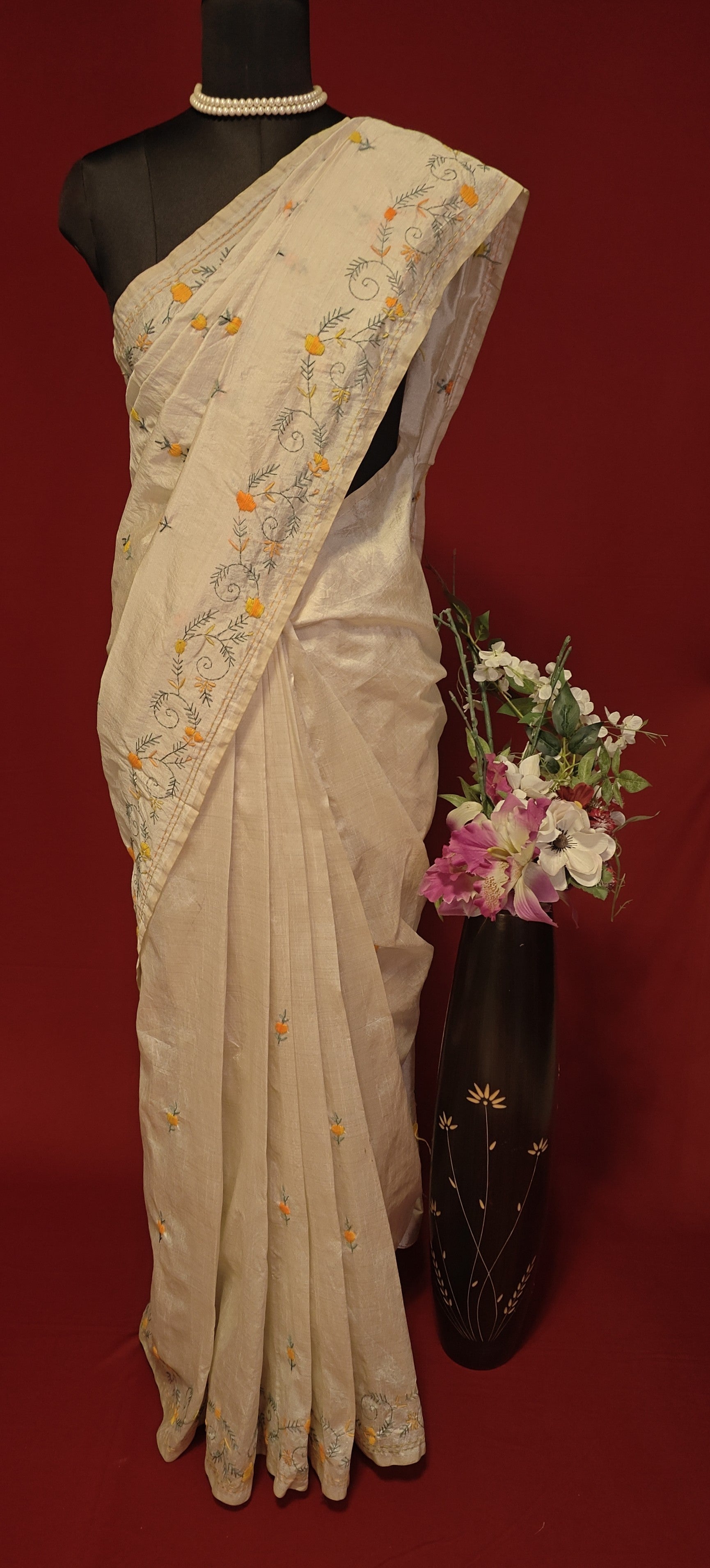 Handcrafted Kantha stitched silk blend saree in cream with multicolor floral and geometric embroidery on a mannequin with flower vase against a red background.