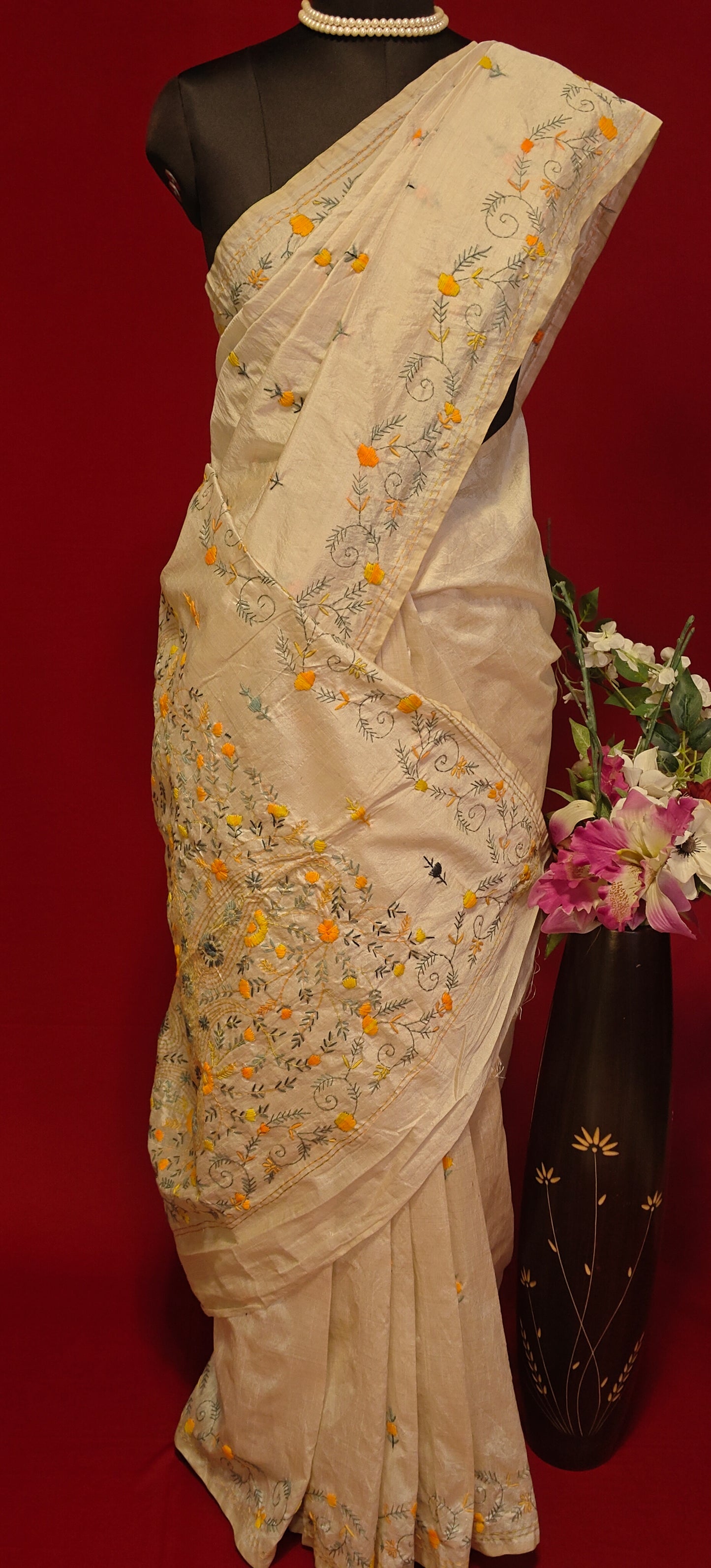 Handcrafted Kantha stitched silk blend saree in cream with multicolor floral and geometric embroidery on a mannequin showcasing the saree pallu with flower vase against a red background.