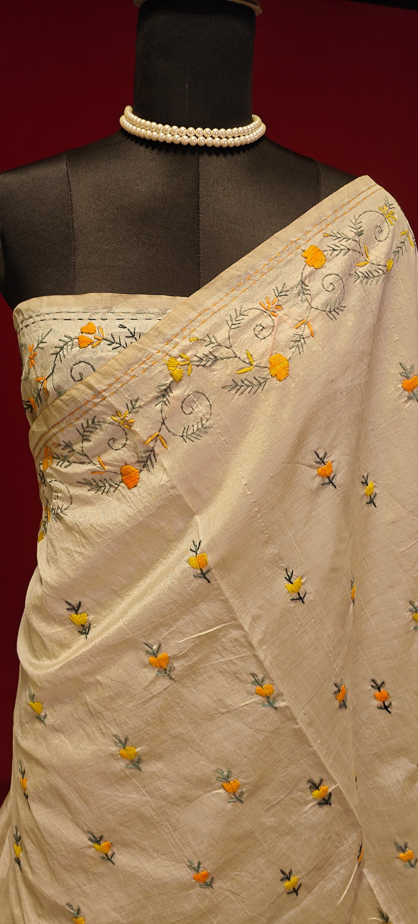Handcrafted Kantha stitched silk blend saree in cream with multicolor floral and geometric embroidery on a mannequin showcasing the blouse piece a red background.