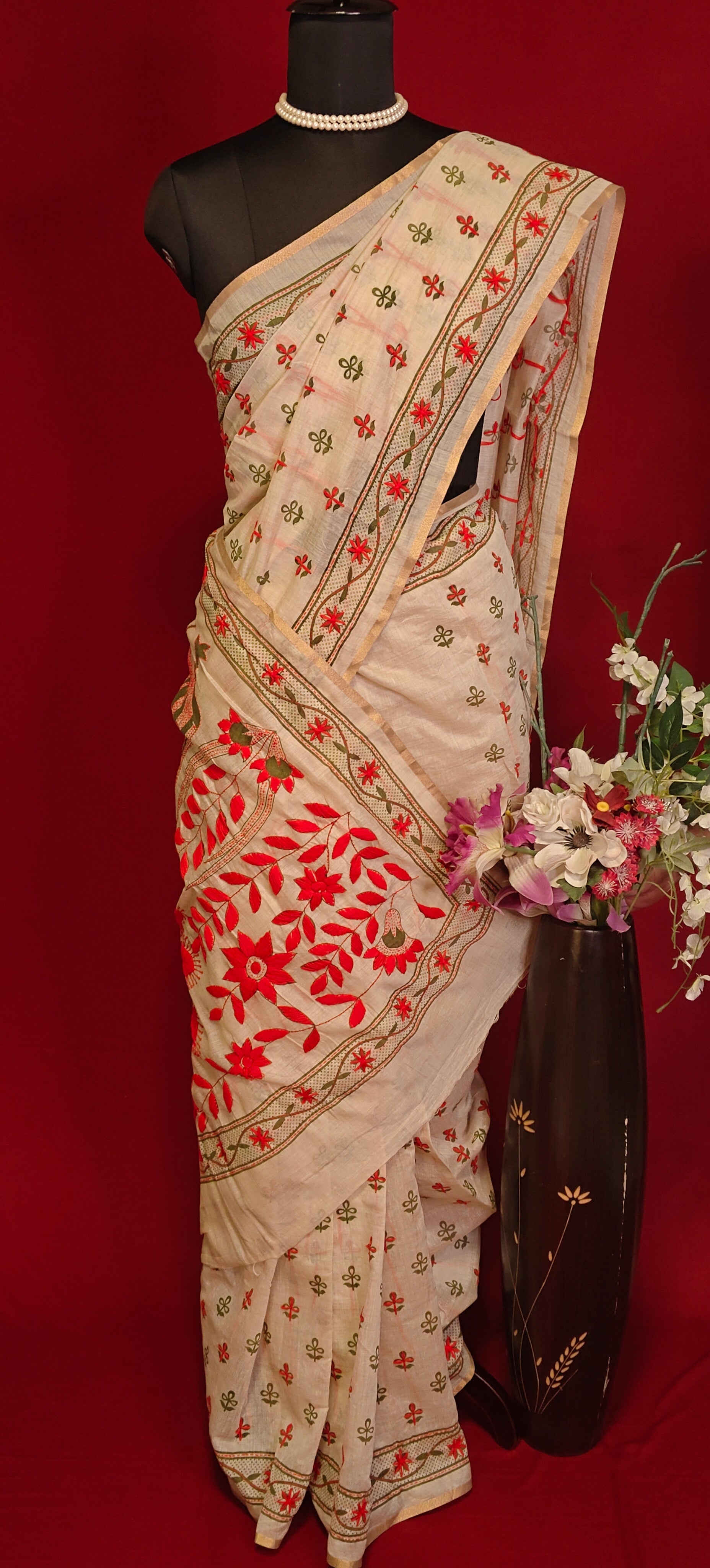 Handcrafted Kantha stitched silk blend saree in cream with red and green floral embroidery on a mannequin showcasing the saree pallu with flower vase against a red background.