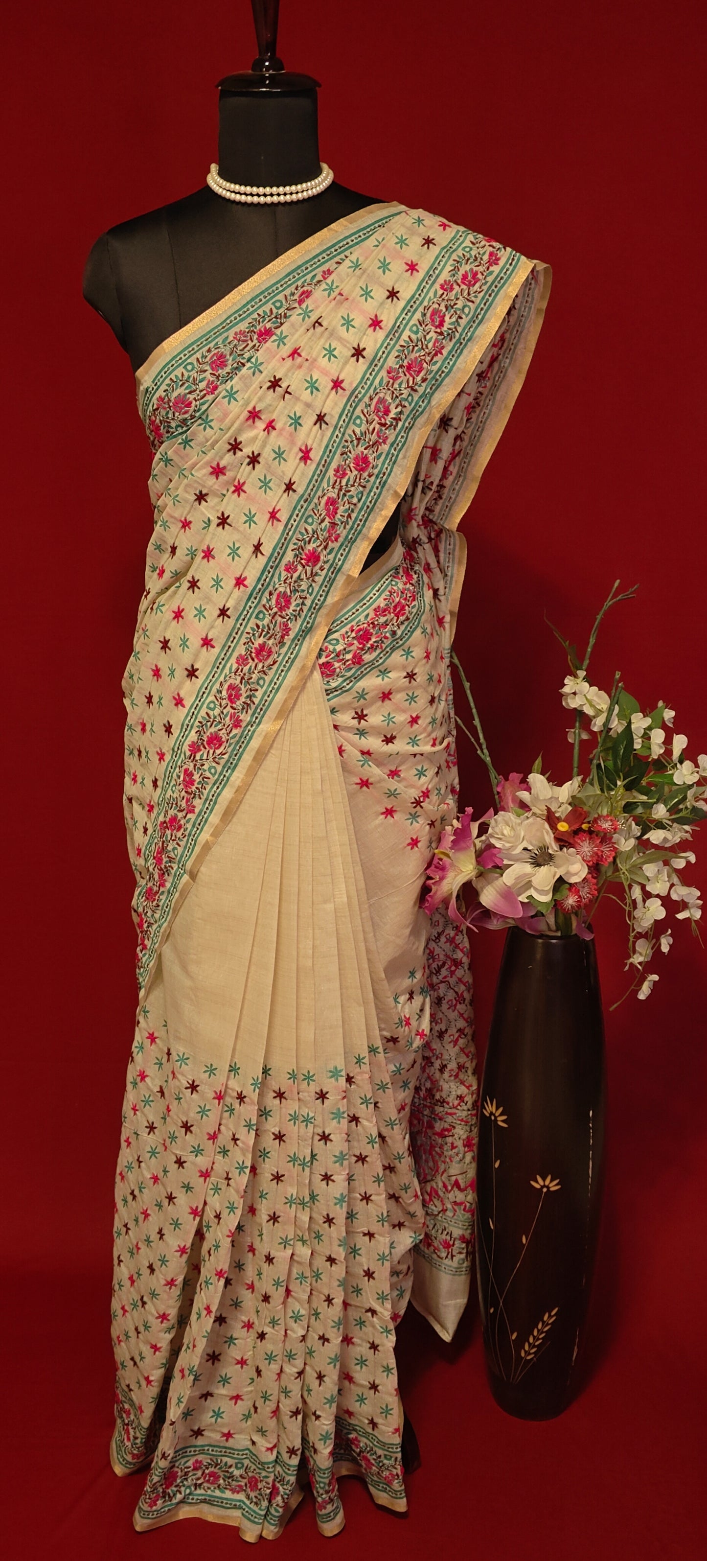 Handcrafted Kantha stitched silk blend saree in cream with pink and sea green floral embroidery on a mannequin with flower vase against a red background.