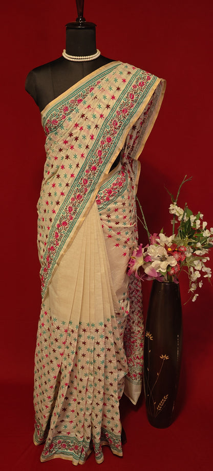 Handcrafted Kantha stitched silk blend saree in cream with pink and sea green floral embroidery on a mannequin with flower vase against a red background.
