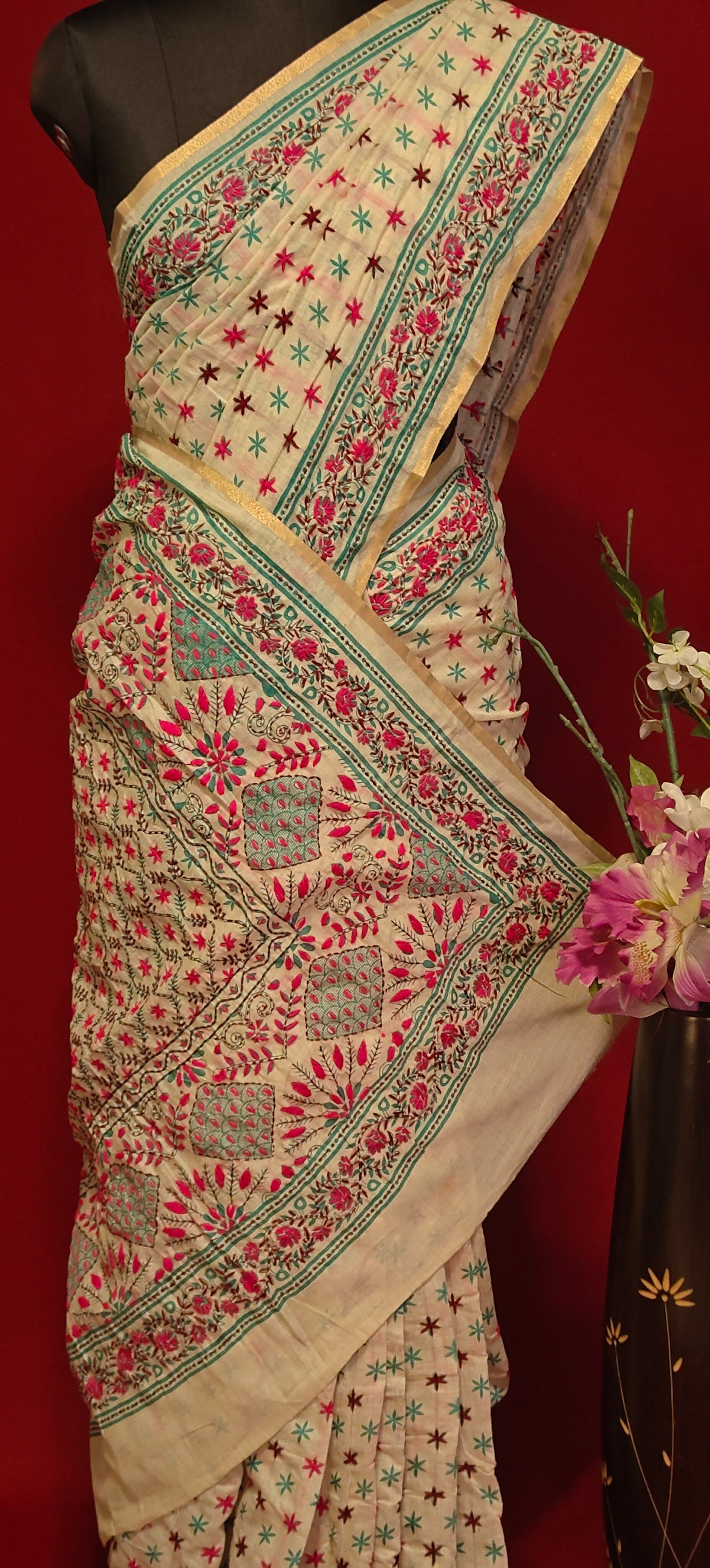 Handcrafted Kantha stitched silk blend saree in cream with pink and sea green floral embroidery on a mannequin showcasing the saree pallu with flower vase against a red background.