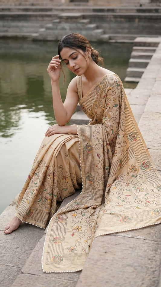 Woman in a handcrafted kantha-stitched cotton saree – gold with multicolor floral & paisley embroidery sitting by a body of water.