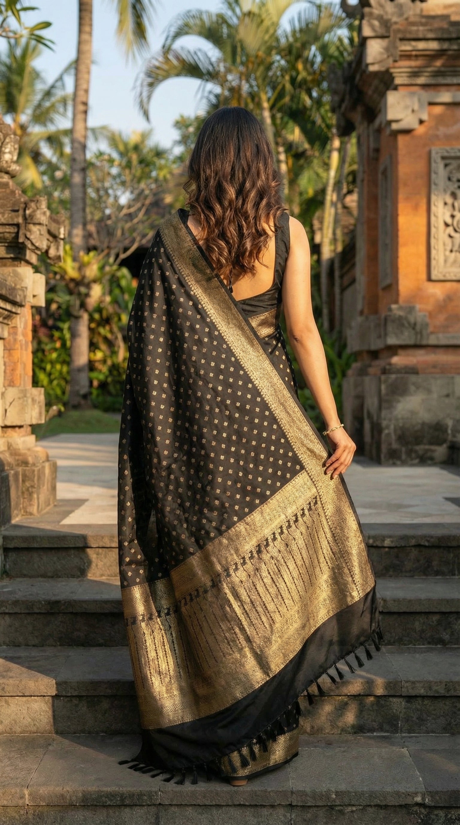 Woman in a Black blended silk saree with silver Khaddi Butti motifs and rich border, standing on stone steps with a temple and palm trees in the background.