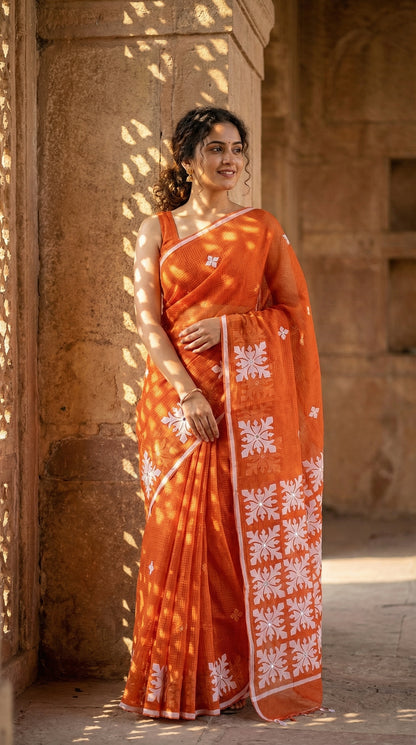 Woman in an orange Kota saree with symmetrical white floral appliqué and soft texture, handcrafted traditional Indian saree standing against a stone wall.