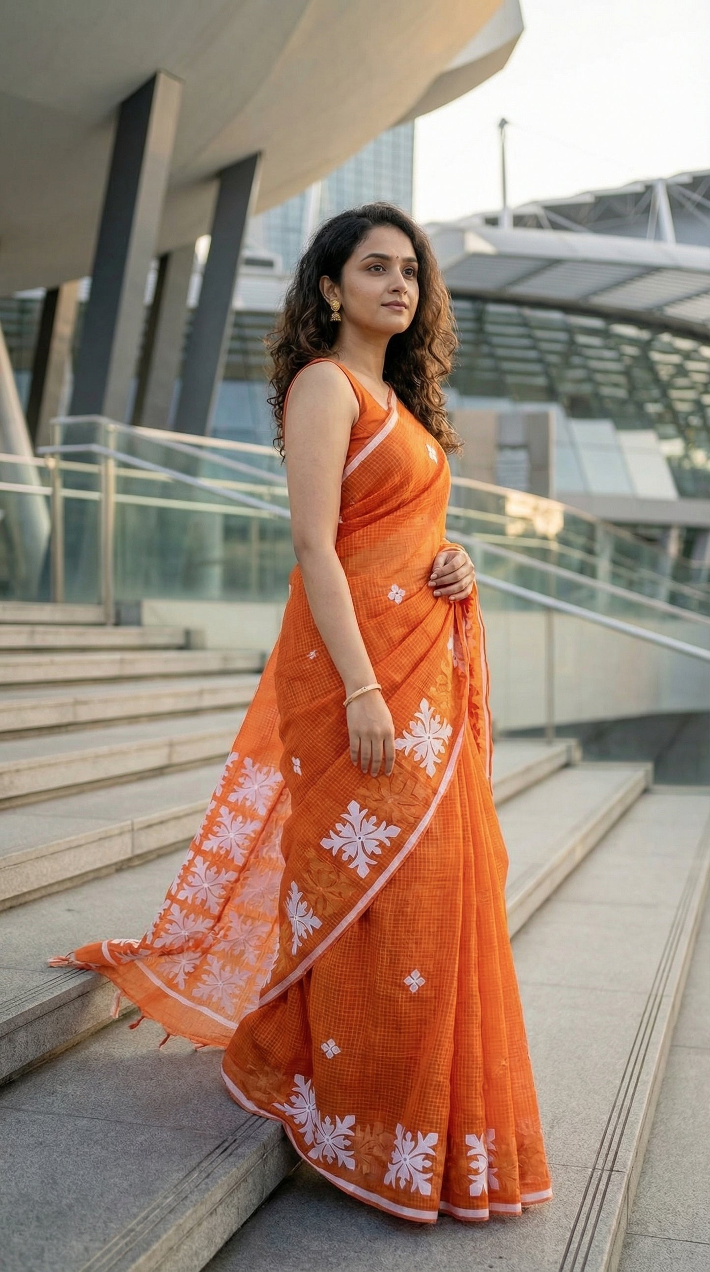 Woman in an orange Kota saree with symmetrical white floral appliqué and soft texture, handcrafted traditional Indian saree standing on steps outdoors.