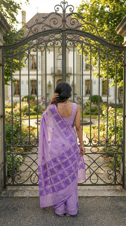 Woman in a lavender-purple-applique-kota-saree, standing in front of an ornate metal gate with a garden and building in the background.