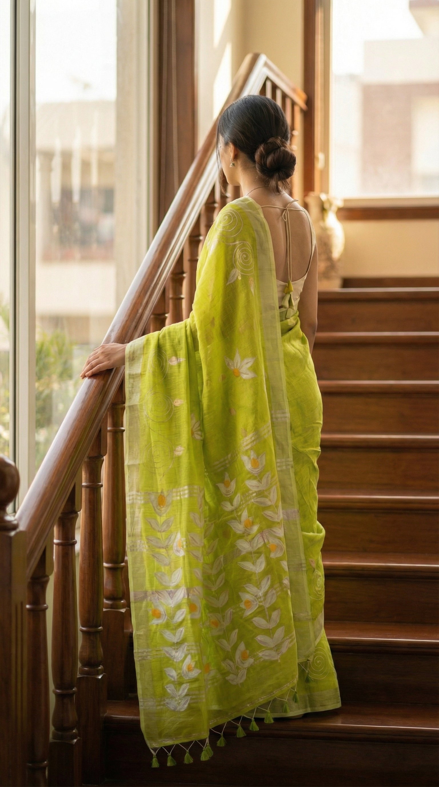 Woman in a lemon green hand painted cotton linen saree with floral motifs and tassel pallu, standing on a staircase.