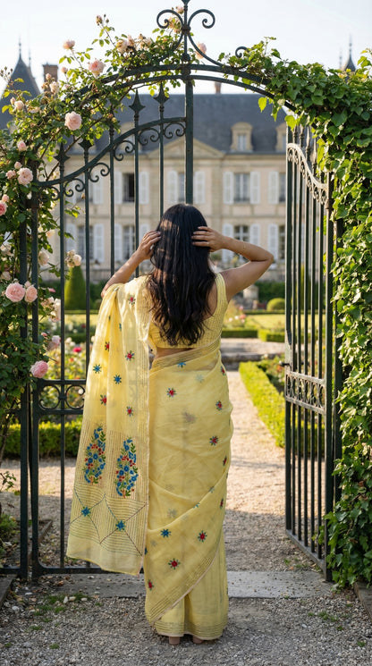 Woman in a lemon-yellow-chanderi-kantha-saree, standing in front of an ornate gate with garden and building in the background