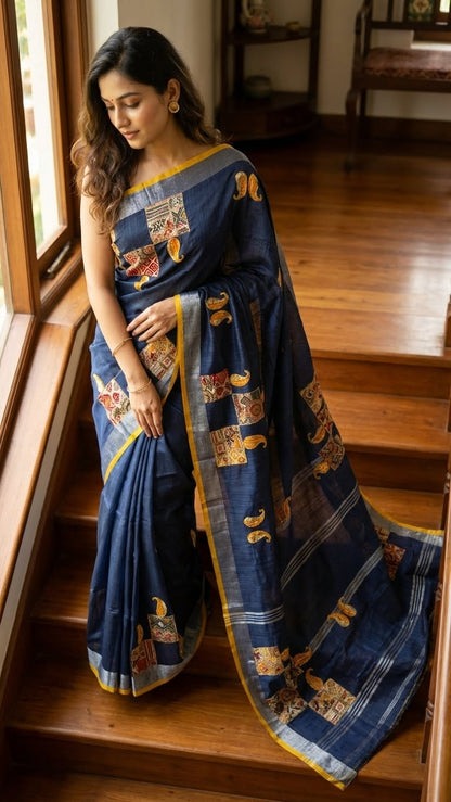 Woman in a grey linen cotton saree with applique work paisley motifs and silver thread detailing standing on wooden stairs.