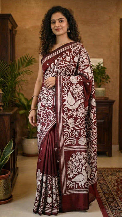 Woman wearing a luxury kantha-stitched silk saree – red with intricate peacock & floral motifs in a home setting.
