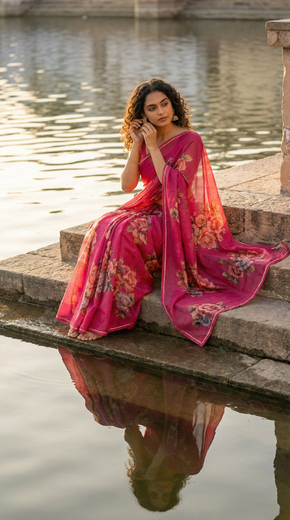 Woman in a magenta chiffon saree with floral prints, sitting by a water body.