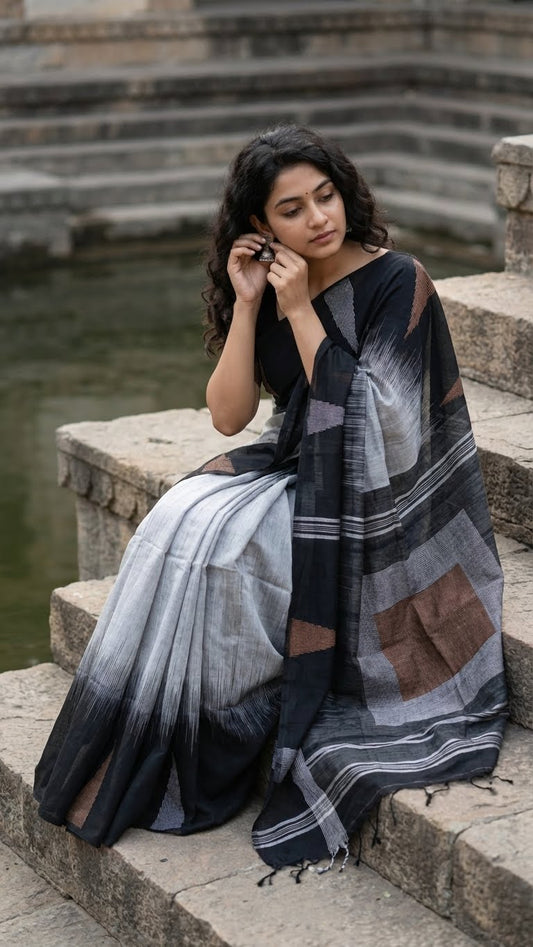 Woman sitting on stone steps wearing a metallic grey blended cotton handloom saree with a bold black border and geometric rust accents by a water body.
