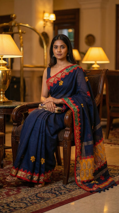Woman in a midnight indigo linen cotton saree with handcrafted applique border, sitting in an elegant room with lamps and decor.