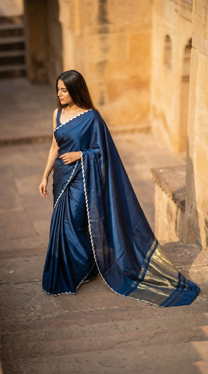Woman in a blue Modal Silk Saree with Lagdi Patta Pallu saree with white border, standing in front of a stone wall.