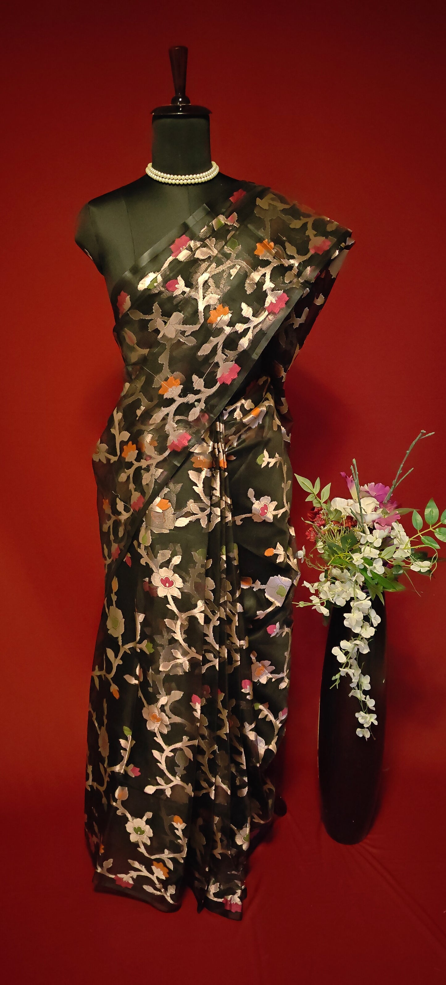 Black muslin blended silk saree with Jamdani weave and gold zari jaal, styled on mannequin with flower vase and red background.
