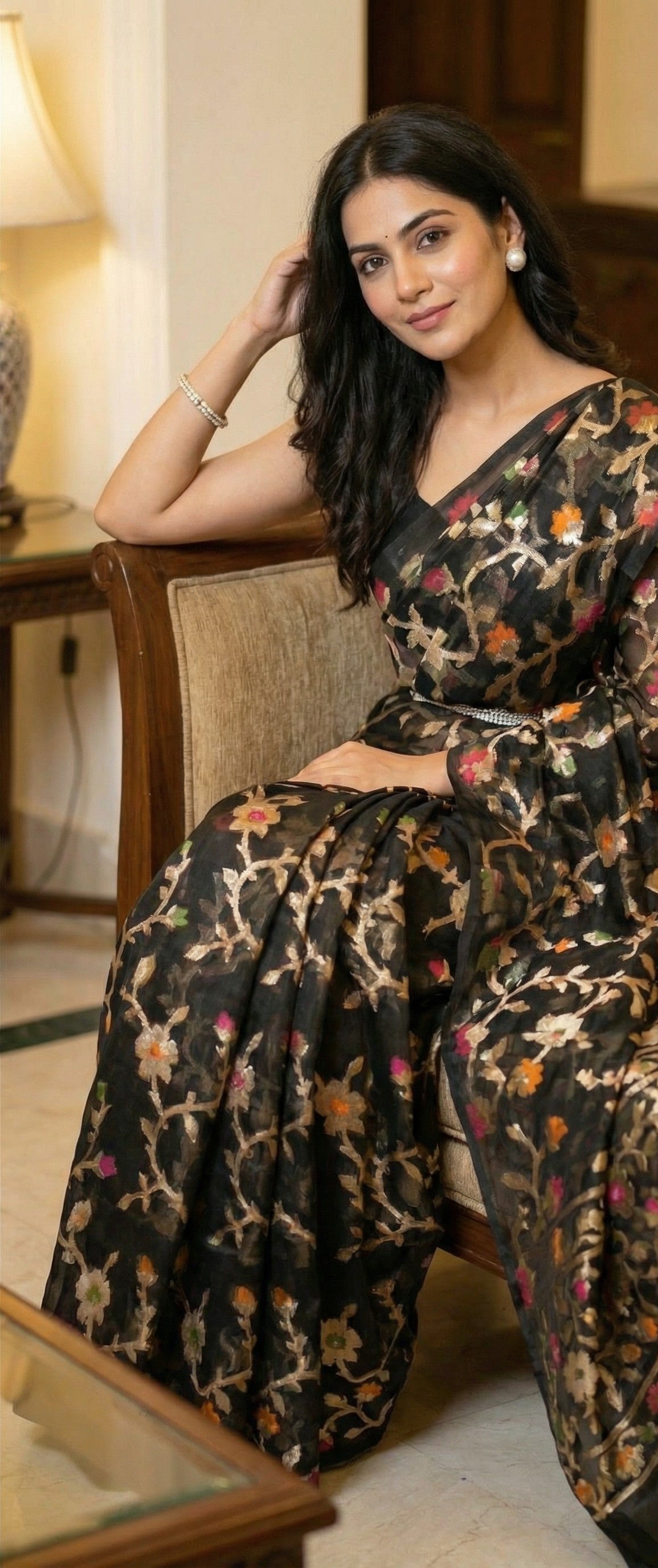 Woman wearing Black muslin blended silk saree with Jamdani weave and gold zari jaal. Sitting on a sofa, leaning back slightly, one arm over the backrest; effortless luxury, blouse + pleats visible.