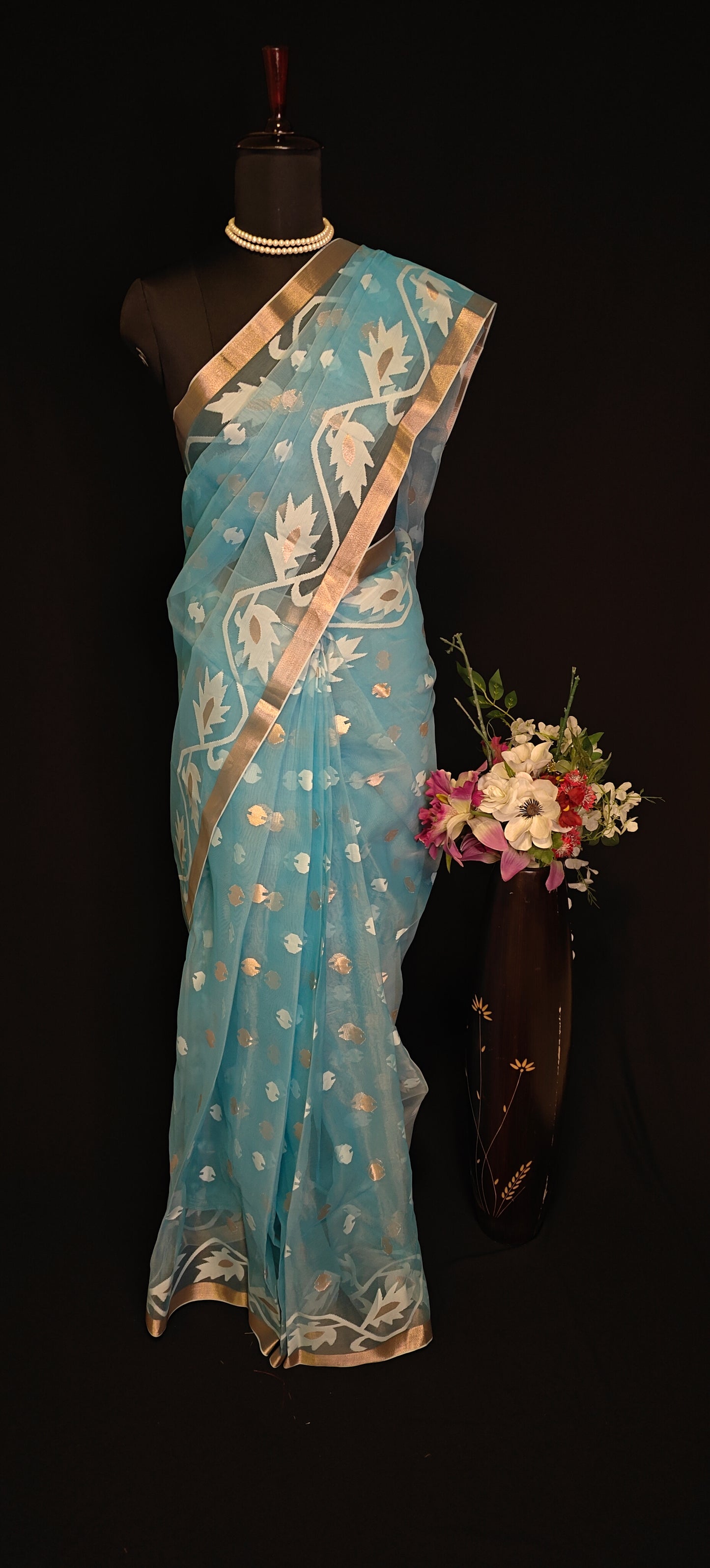 Sky blue organza muslin saree with golden border and Jamdani floral embroidery, styled on mannequin with flower vase against a black background.