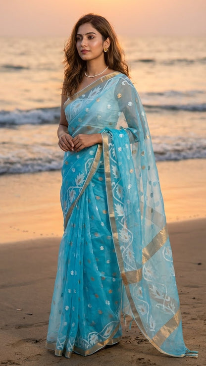 Woman in a sky blue organza muslin saree with golden border and Jamdani floral embroidery standing on a beach at sunset