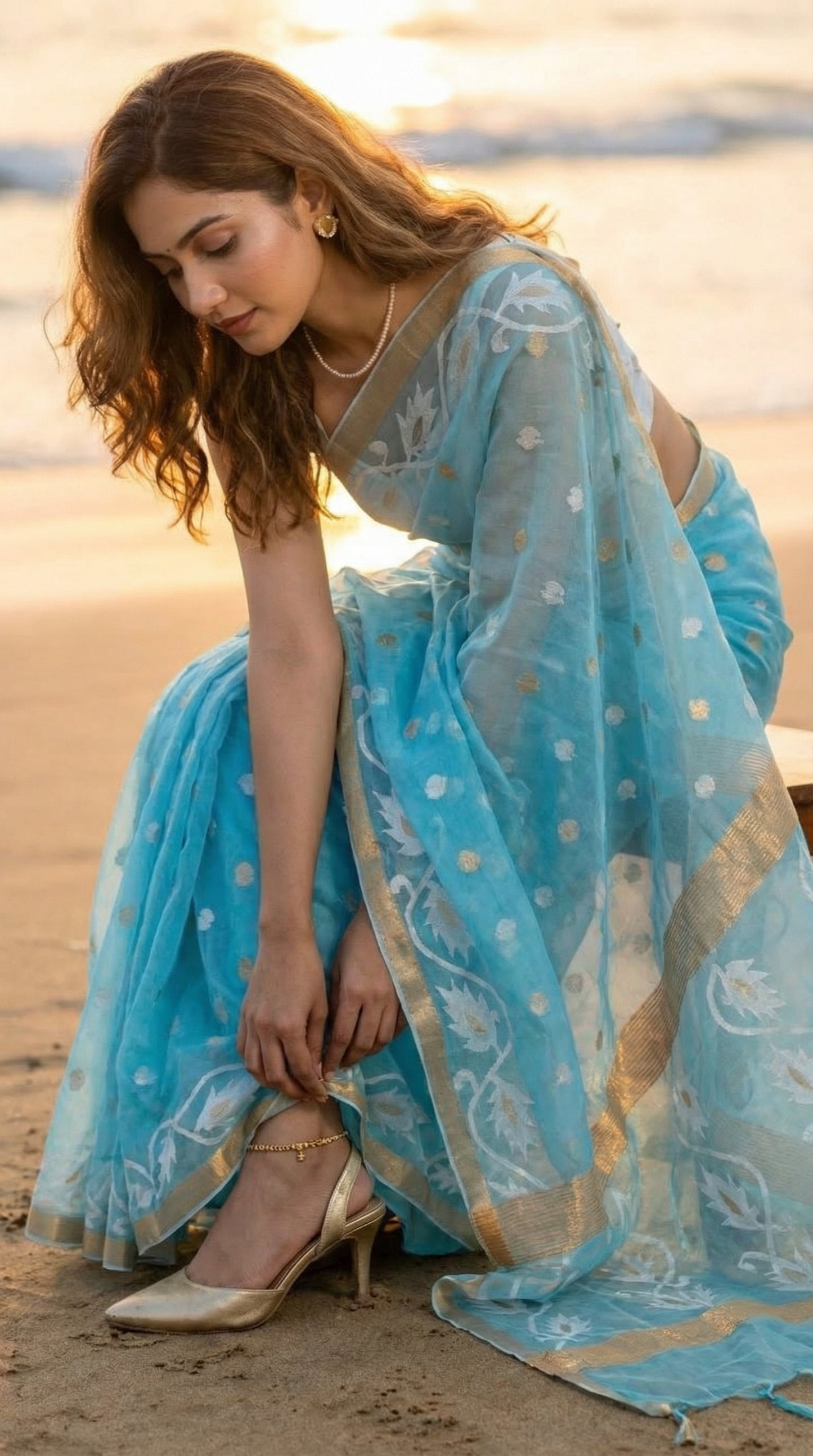 Woman in a sky blue organza muslin saree with golden border and Jamdani floral embroidery, sitting on a beach