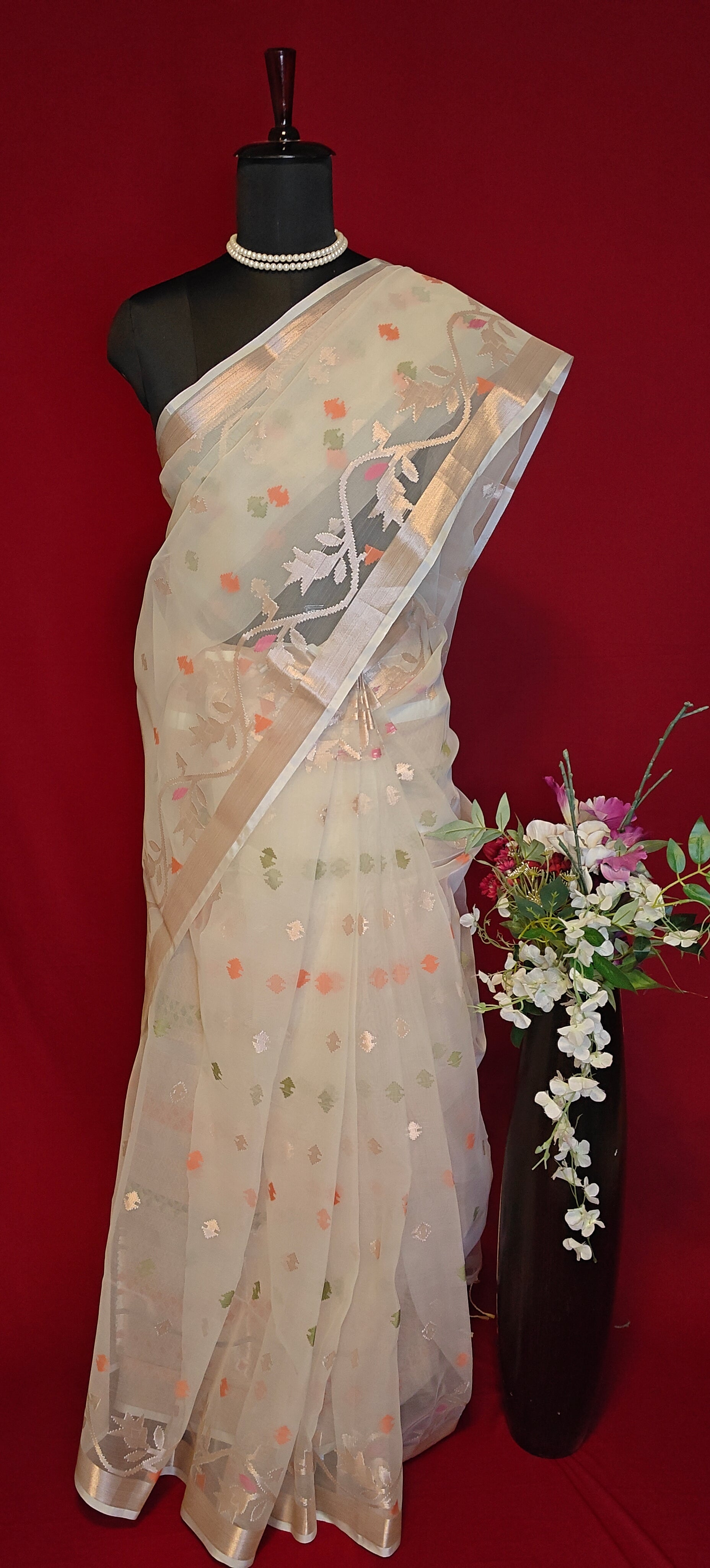 Cream muslin blended silk saree with golden border and pastel Jamdani motifs, styled on mannequin with flower vase against a red background.