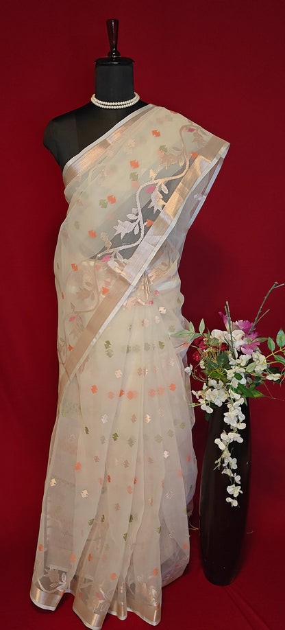 Cream muslin blended silk saree with golden border and pastel Jamdani motifs, styled on mannequin with flower vase against a red background.