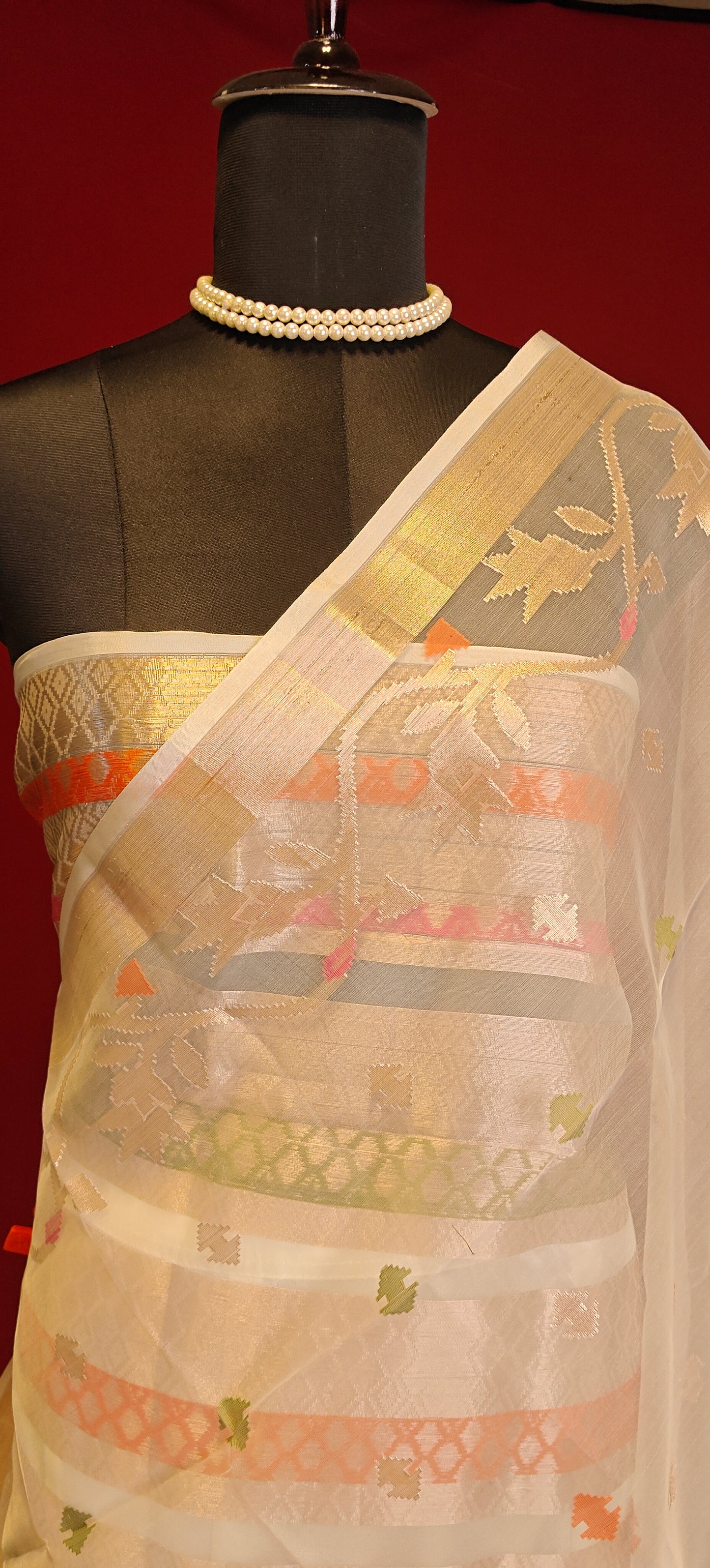 Cream muslin blended silk saree with golden border and pastel Jamdani motifs, styled on mannequin showcasing the blouse piece against a red background.