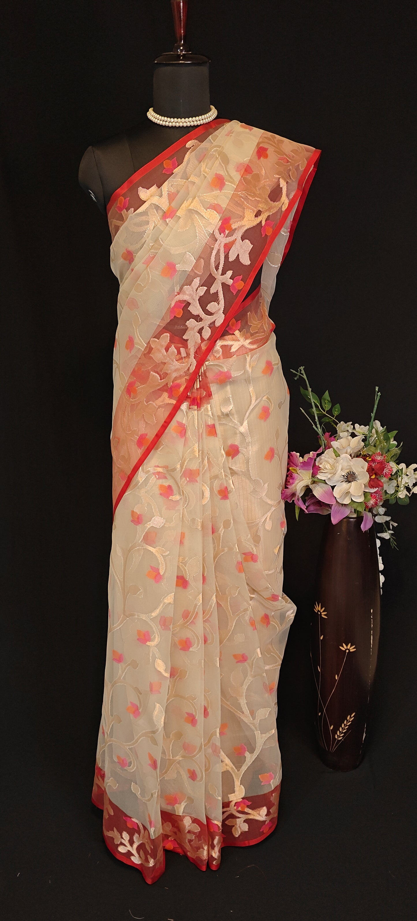 Cream muslin organza saree with red border and Jamdani floral embroidery, styled on mannequin with a flower vase and black background.
