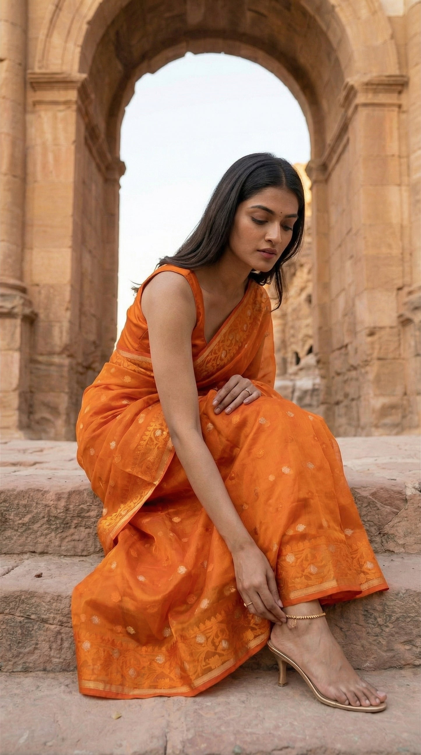 Woman in an orange organza muslin saree with Jamdani weave and gold-silver Zari booti, sitting on steps with a stone archway in the background.