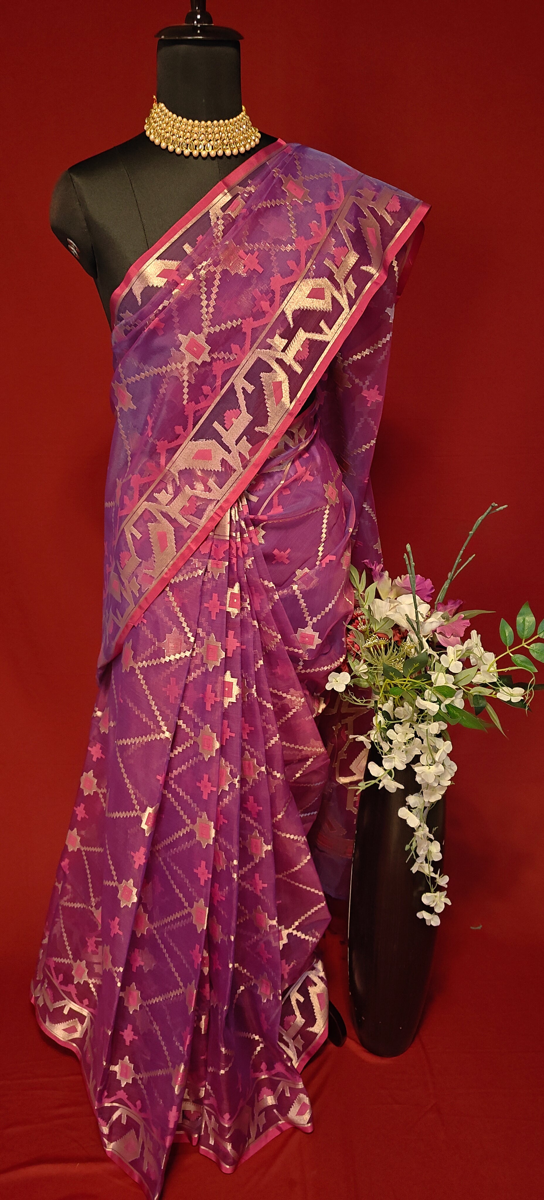 Purple muslin silk saree with Jamdani weave and golden motifs, styled on mannequin with a flower vase against a red background.