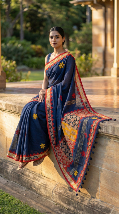Woman in a navy blue linen cotton saree with traditional applique border, sitting on a stone ledge outdoors.