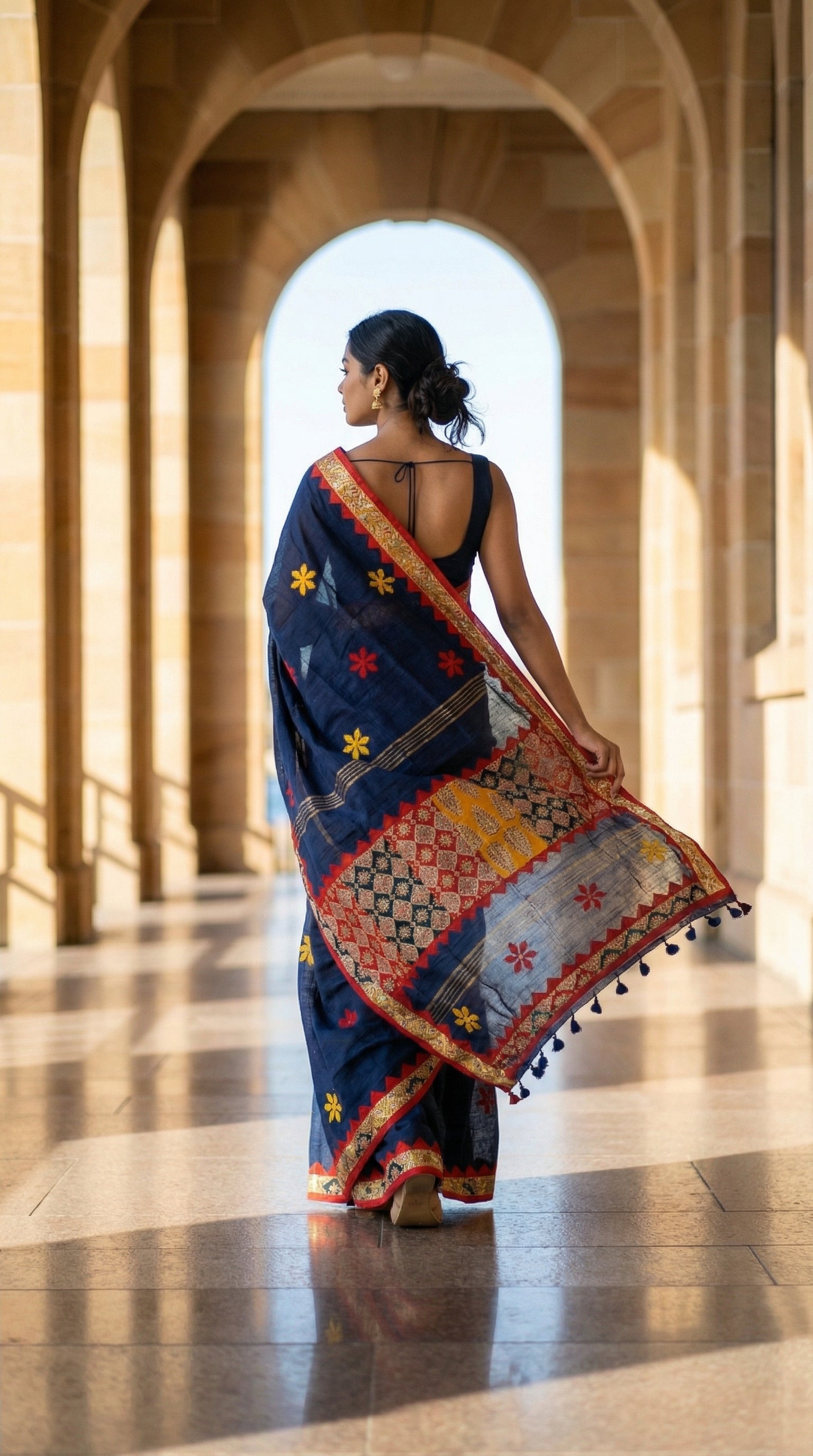 Woman in a navy blue linen cotton saree with traditional applique border, walking through a sunlit architectural hallway.