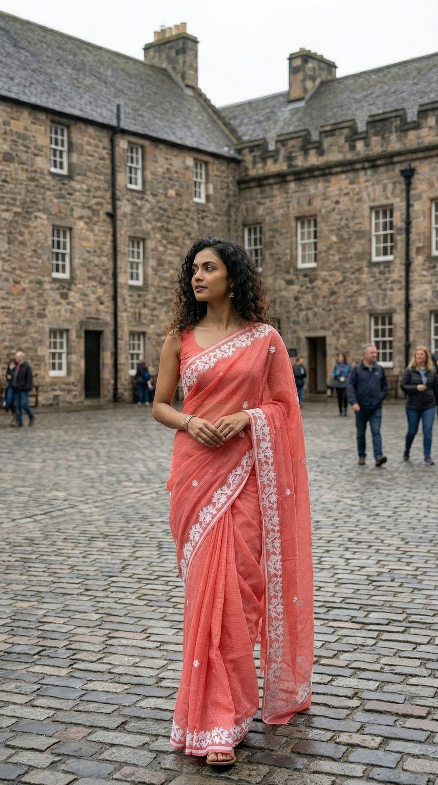 Woman in a peach-kota-doria-saree, standing on a stone pavement with a historic building in the background.