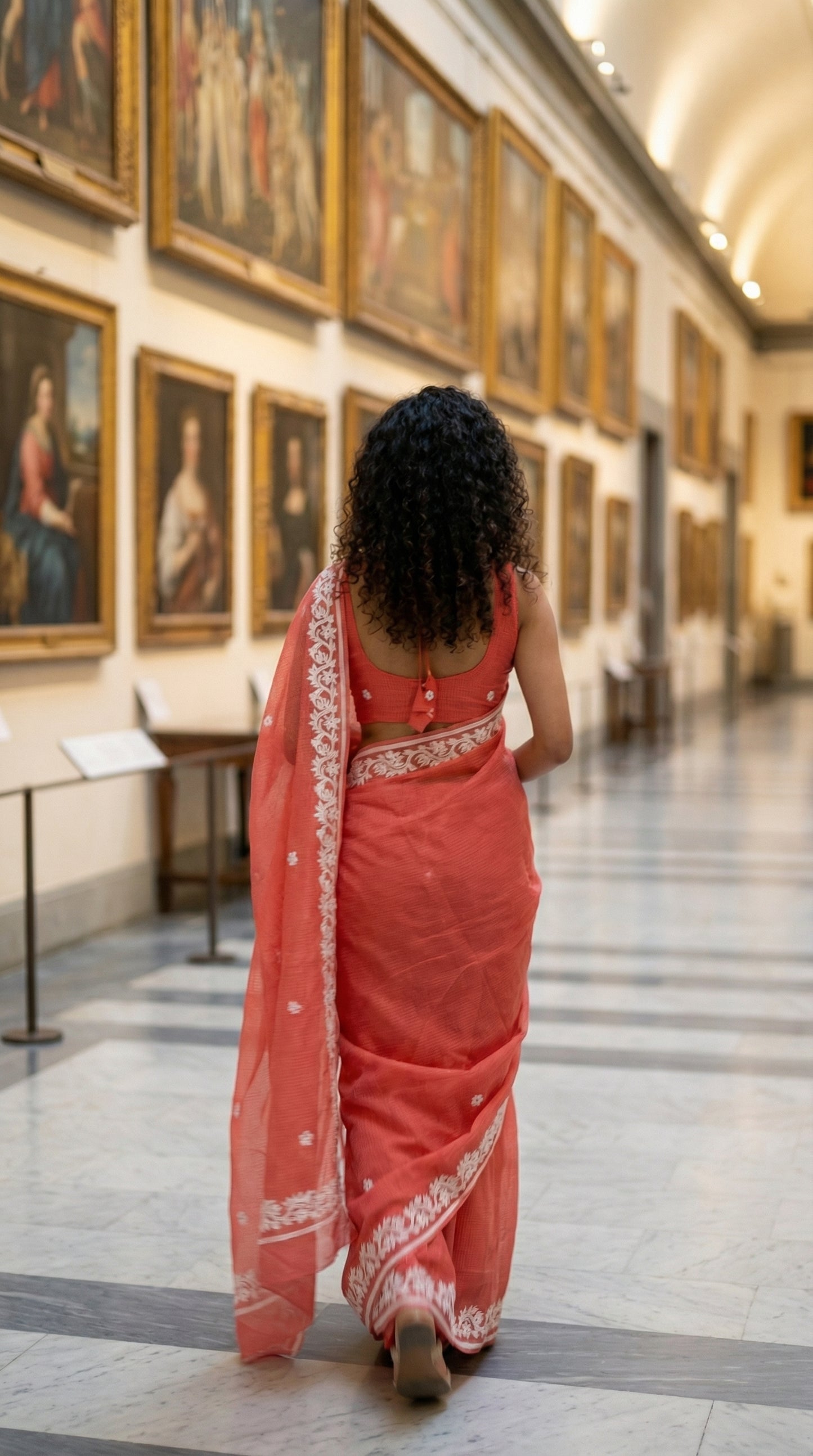 Woman in a peach-kota-doria-saree, walking through a museum with paintings on the walls.