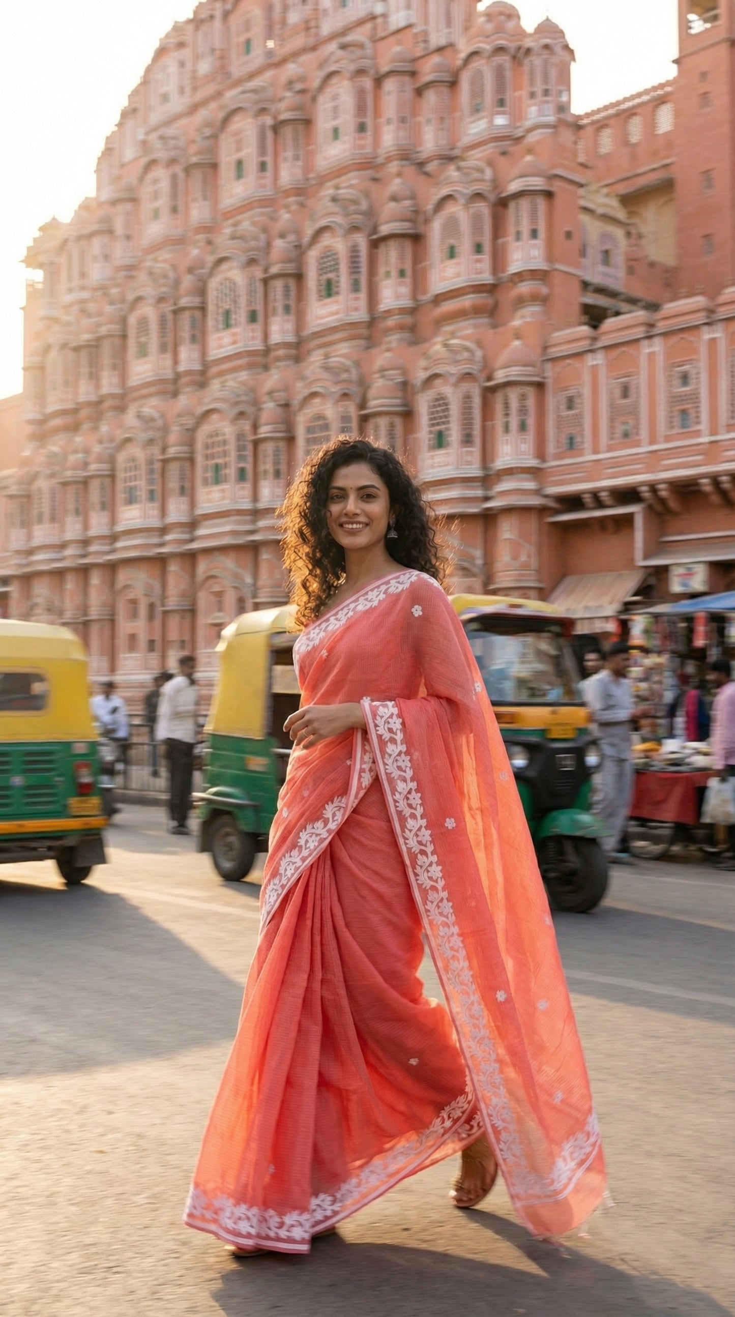 Woman in a peach-kota-doria-saree, walking in front of a historic building with auto-rickshaws in the background.