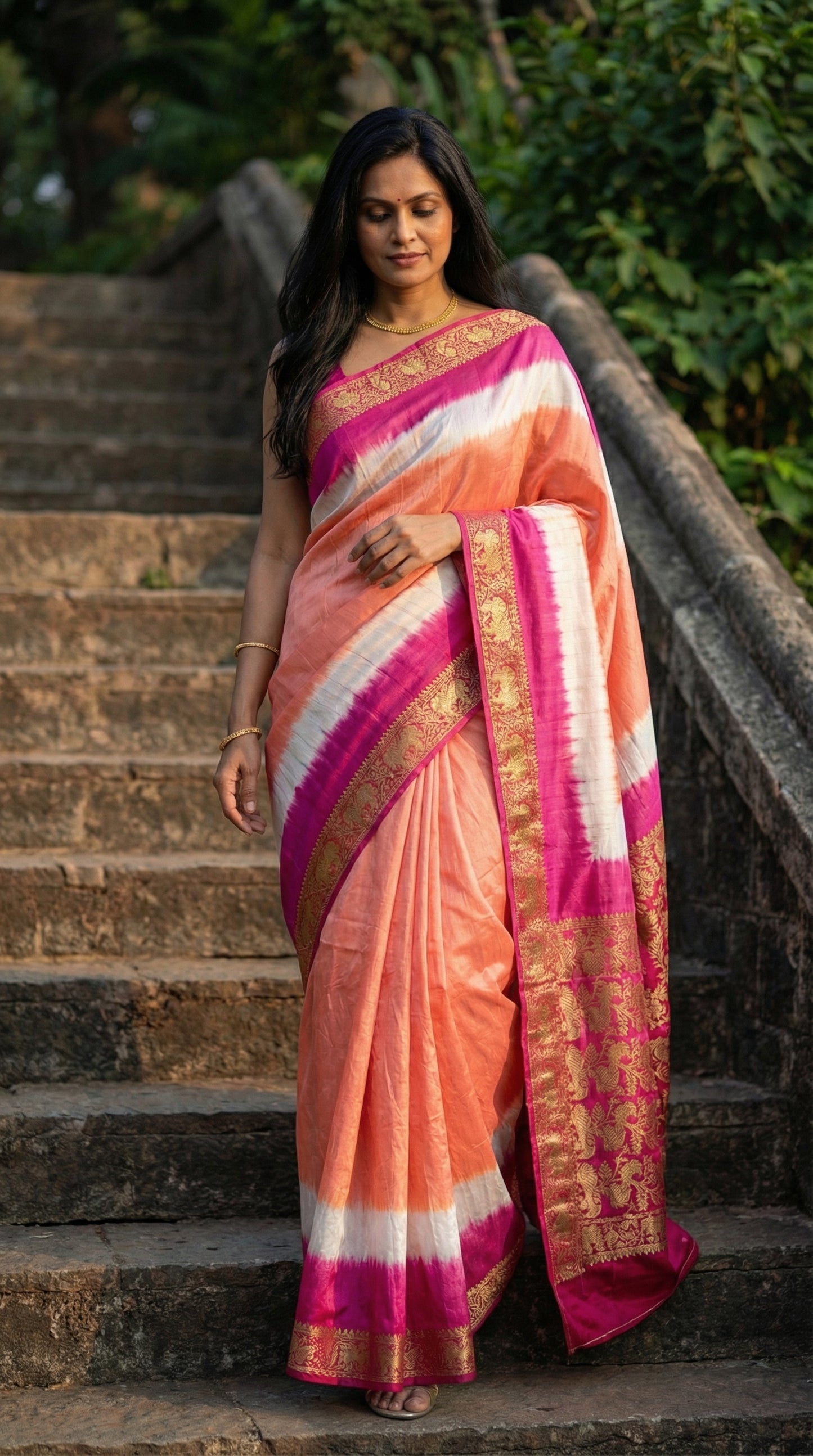 Woman in peach modal silk saree with pink Zari border and brocade pallu, with pink and white stripes on stone steps.