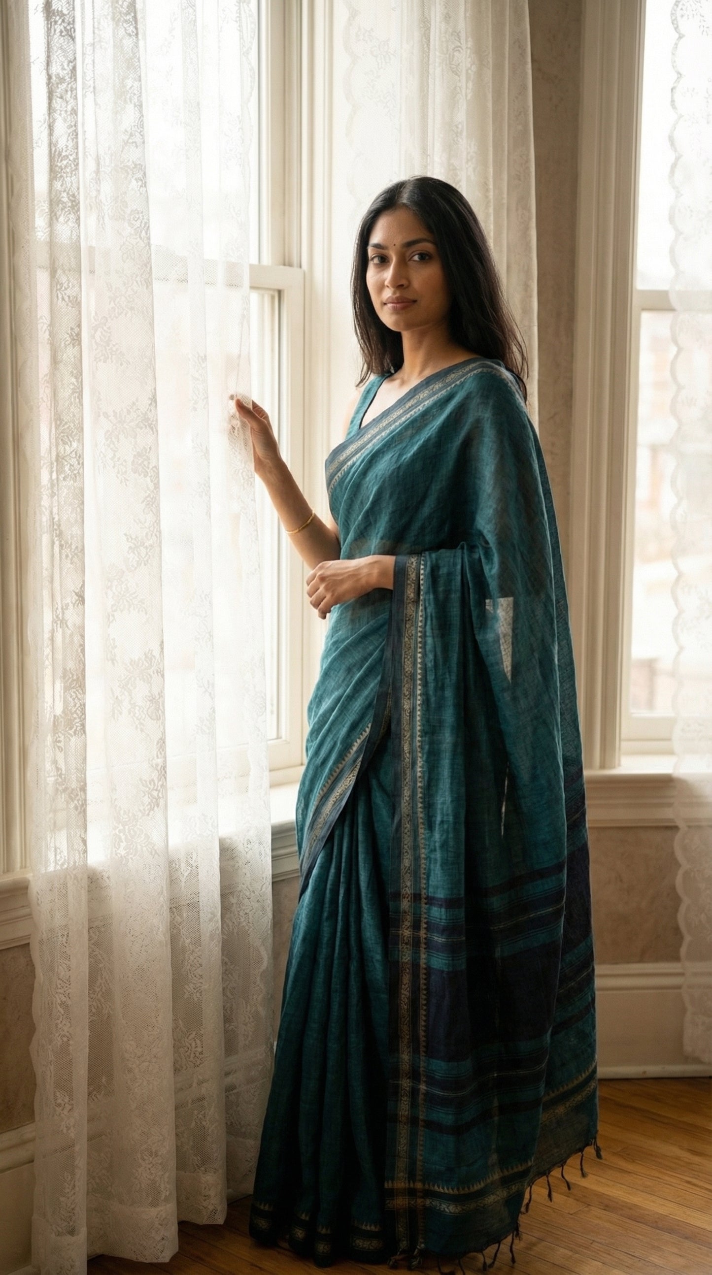 Woman wearing a peacock-green-mul-cotton-saree, standing in a room with large windows.