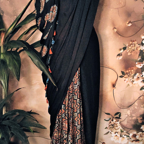 Ajrakh Modal Silk Saree – Black with Earthy Motifs and Tassel-Embellished Pallu saree with floral patterns on a decorative background.
