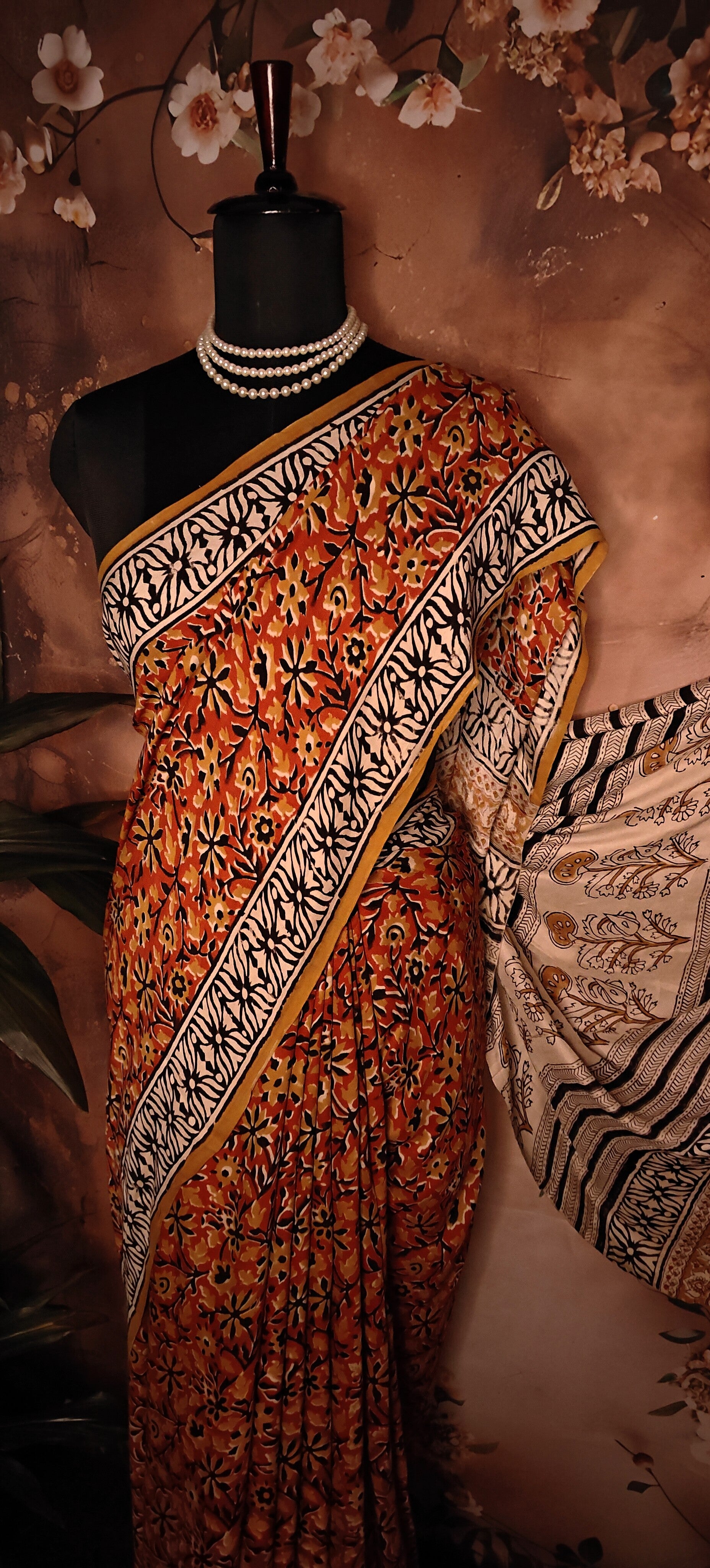 Ajrakh Modal Silk Saree – Reddish-Orange with Black & White Border and Golden Pallu Motifs saree with intricate patterns on a mannequin against a floral background