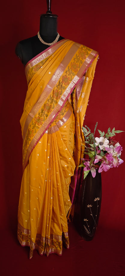Yellow blended silk saree with shimmering zari border, styled on mannequin with a floral arrangement and a dark background.