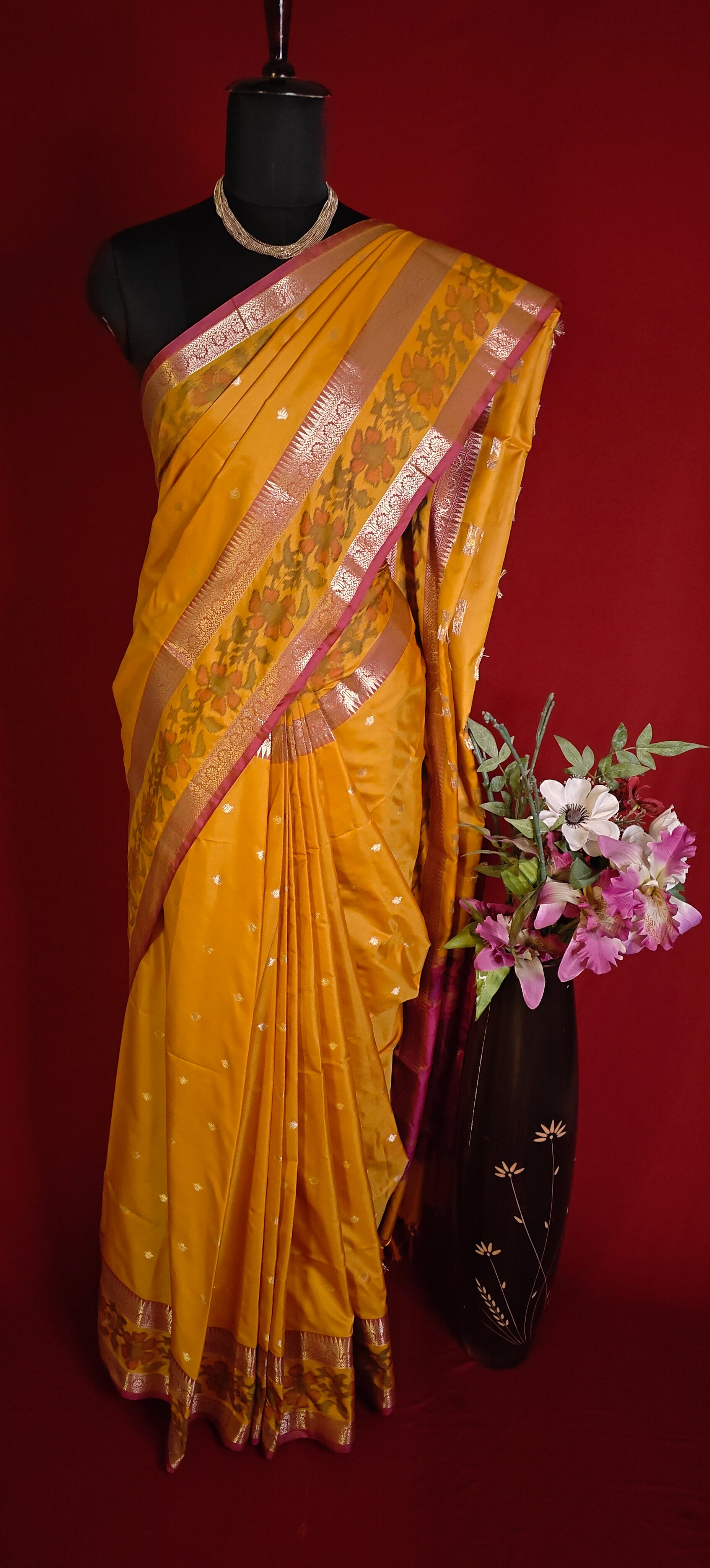 Yellow blended silk saree with shimmering zari border, styled on mannequin with a floral arrangement and a dark background.