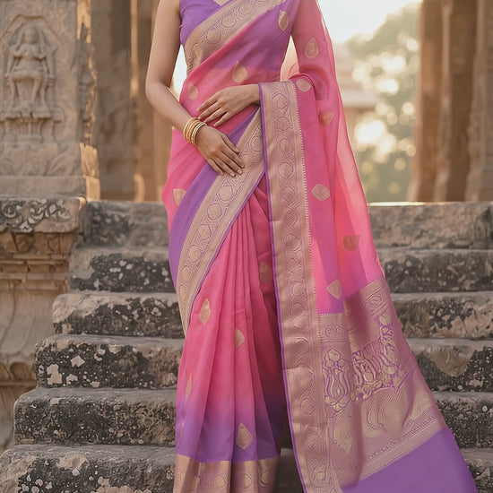 A front portrait of a woman walking down the stone stairs gracefully, showcasing the sheer organza texturing, gold buttis, and intricate purple zari paneling of her pink Banarasi silk saree