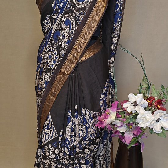 Black silk saree with Kalamkari-style floral and paisley print and golden border on a mannequin wearing a pearl necklace, against a plain background.
