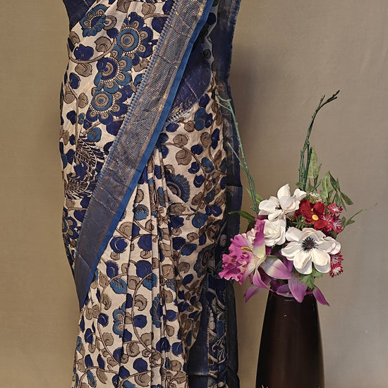 Indigo silk saree with Kalamkari-inspired digital print and golden border on a mannequin, next to a vase with flowers on a plain background.