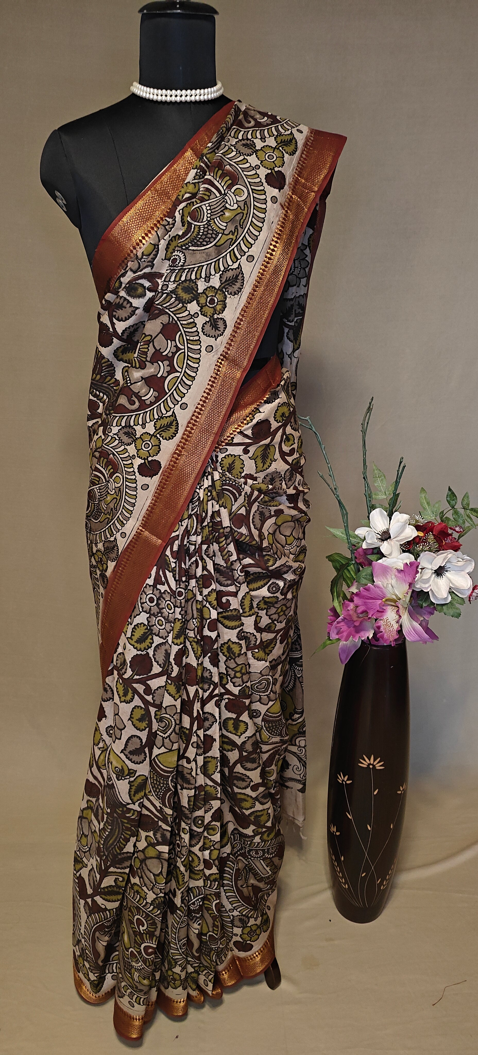 Brown silk saree with Kalamkari-inspired motifs and subtle golden border on a mannequin wearing a pearl necklace, against a neutral background.